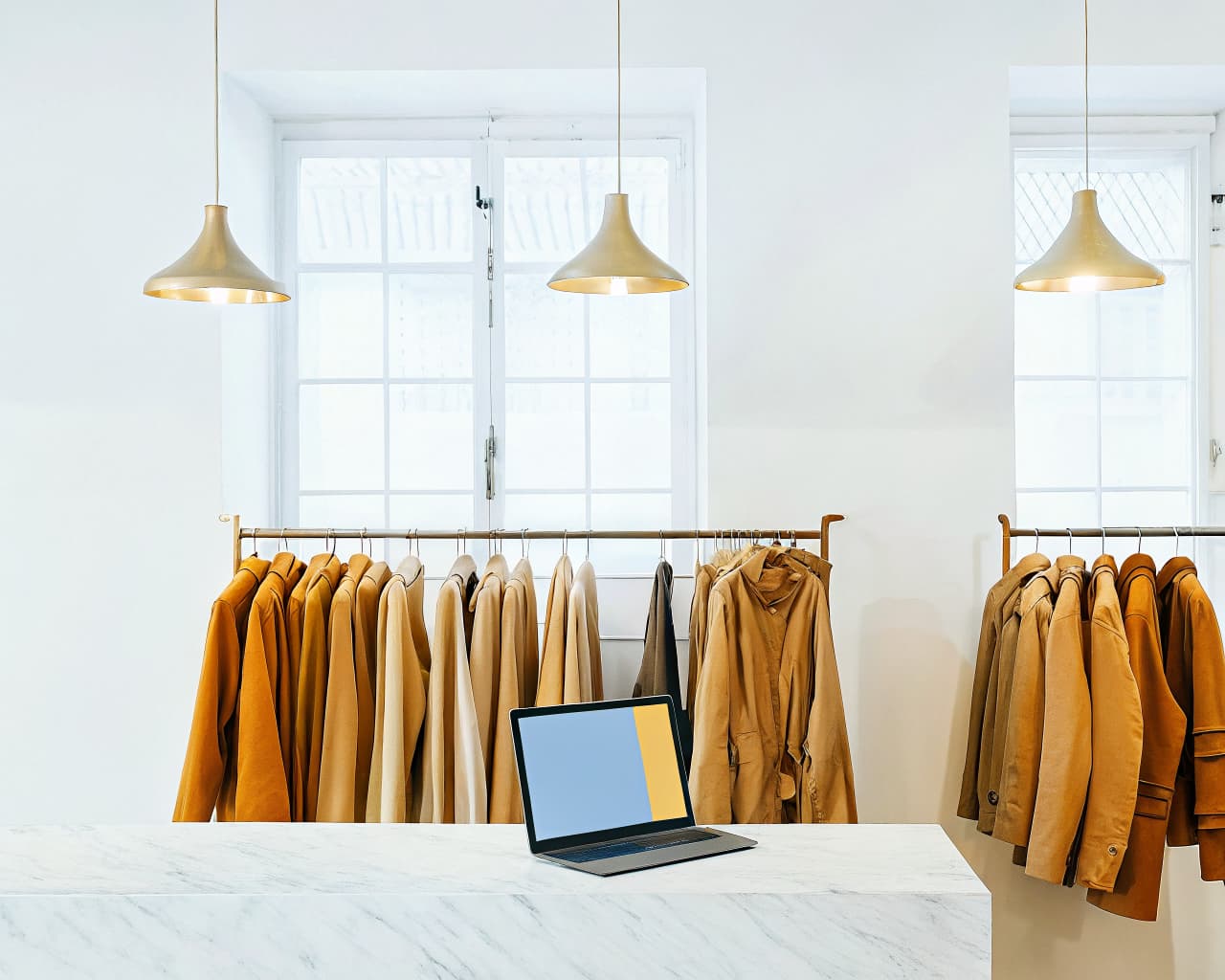 Bright boutique clothing shop interior with racks of jackets and a laptop displaying a colorful product page on a marble counter
