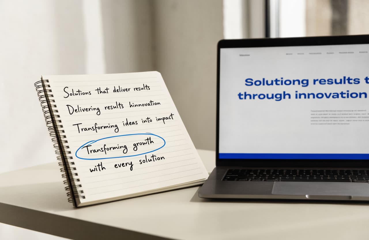 Notebook on a desk with several handwritten headline options, one circled in blue pen, next to a laptop showing a website hero section